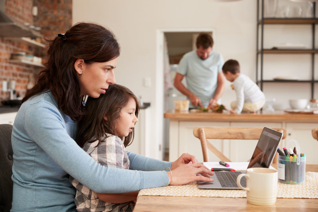 Image of a family in the kitchen. In the foreground there is a mother and son looking at a laptop. In the background there is a father and son cooking a meal.