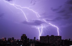 Image of a lighting storm in a city. Buildings are in the foreground and a streak of lighting fills the sky in the background.