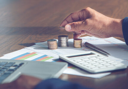 Man counting coins on a pile of financial papers. There is a pen, calculator and laptop on the papers as well.